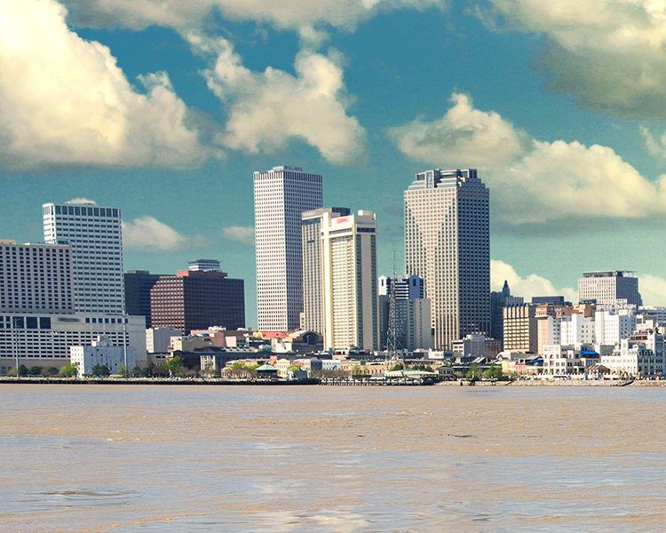 Skyline of New Orleans with various high-rise buildings, viewed from across a wide river under a partly cloudy sky.